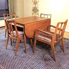 Mid Century Modern Dining Table and Chairs by Mount Airy Chair Co: A Mid Century Modern teak dining room table and chairs from Mount Airy Chair Co. This dining room set features a table with a rectangular surface. The table features side drop down leaves and one insert leaf. The table top rests on four tapered legs and is labeled with a tag from Mount Airy Chair Company. The chairs feature a curved crest rail and slanted, open back leading to an upholstered seat supported by four tapered legs.