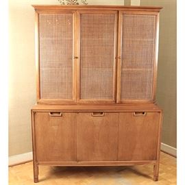 Mid Century Modern "Janus" Cabinet by Mount Airy Furniture: A Mid Century Modern china cabinet, designed by John Stuart for Mount Airy Furniture’s Janus Collection. The cabinet is two pieces and features two cabinets in the upper portion leading to two shelves. The lower portion features two cabinet doors opening to a shelf and a cabinet door opening to three pullout drawers. The cabinet is composed of wood and features details of cornice molding and woven cane. The inside drawer is marked, “Mount Airy Janus Collection.”