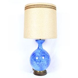 Large Mid Century Modern Ceramic Lamp: A large Mid Century Modern ceramic lamp. This lamp features a fluted and narrow neck leading to a rounded form. The ceramic body of lamp features a swirled blue finish. The body of the lamp sits on a wooden and gold tone base. Included with the lamp is a lamp is a large barrel shade consisting of cream fabric with gold and brown detail. The lamp features two light sockets and can be operated with a pull chain.