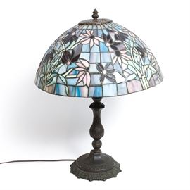 Tiffany-Inspired Table Lamp: A Tiffany-inspired table lamp. This lamp features a stained glass shade with a floral motif expressed in hues of blue and purple. It has a curved column and an ornately decorated base. This lamp is unmarked. It is tested and operable.