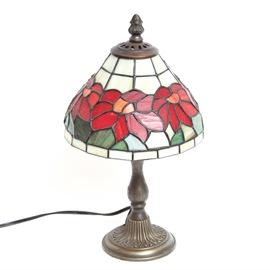 Tiffany-Inspired Table Lamp: A Tiffany-inspired table lamp. This lamp features a stained glass shade with a red floral motif. It has a curved column and rests on a round base with a flower petal decoration. Item is tested and operable. Unmarked.