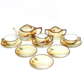 Vintage Handpainted Nippon Tea Set: A vintage handpainted Nippon tea set. This porcelain set features a waterfront scene with palm trees and a building. The collection includes a tea pot, cream, double handle lidded sugar bowl, two cups and six saucers. They are marked, “Handpainted Nippon” to the bottom.