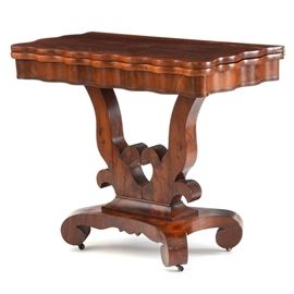 French Empire Style Scallop Game Table: A French Empire style scallop game table with a mahogany finish. It features a scalloped edge and apron and revolves and flips down to convert into a larger table. It rests on a lyre base with a pierced heart motif, scrolled legs, and casters.
