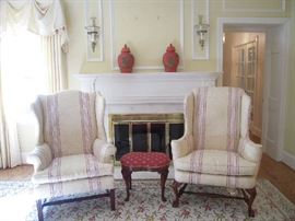 Living Room:  Two ivory/raspberry stripe wingback chairs are almost identical.  The one on the left has Chippendale style legs and rolled arms while the one on the right has claw/ball feet and narrow flat arms. Both were upholstered by LUTTRELL UPHOLSTERING.    Did you know that this style of chair was developed in England in the 1600's where cold weather made the roaring fireplace the gathering spot of the house?  The original style was created to keep women warm as they lounged in the wingback chairs to protect their upper chests from cold drafts. The design gained popularity in the early 1700's and eventually morphed from a wooden chair to one with upholstery.   On the mantel are two red/gold urns with lids; attached to the wall [but easily removable] is a pair of brass/glass candle hurricanes.    