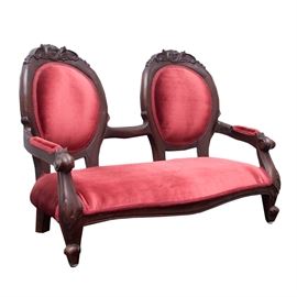 Louis XV Style Double Doll Chair: A Louis XV double doll chair. This chair for dolls features a carved wooden frame with floral and foliate accents at the crest rail, upholstered armrests with claw arms, with foliate accent legs with pad feet. The chair is upholstered with red and features a braided border around the upholstery in a light red.