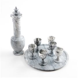 Grey Marble Barware Set: A grey marble barware set. This set offers a decanter with a tiered topper, a tall neck, and a small body. It includes six small stemmed cups and a round tray. Several pieces have decals marked “Made in Taiwan Republic of China”.
