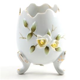 Japanese Porcelain Footed Egg Vase: A Japanese porcelain egg vase. This piece is in the form of a egg shell that has the top portion broken of. It rests on three feet and has applied yellow roses and green leaves with gilt details. The piece is marked with crossing swords in blue and is numbered “55/1144”, and has a green on gold label that reads “Japan”.