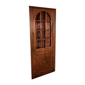 Oak Corner Cabinet: An oak corner cabinet. This piece features an upper door with an arched glass front with muntin. Below is a recessed panel door with an urn applique. The doors are flanked by fluted trim, and open to reveal display and storage shelves within. The piece stands on a plinth base.
