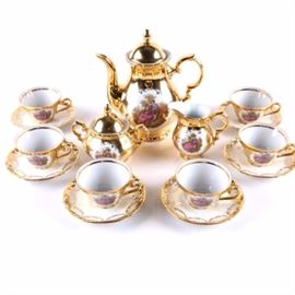 Gloria Porcelain Tea Service: A Gloria Porcelain Company tea service. It include a teapot, cream and covered sugar and six cups and saucers. Each of the pieces, except saucers, is decorated on one side with an image of a man and woman sitting in a wooded area and with bright gold tone gilt covering the rest of the exterior. The saucers have raised decoration along the rims, gilt edges and painted scroll decoration. The pieces are marked to the underside “Gloria Fine Porcelain Bavaria Handwork Bayreuth West-Germany”.
