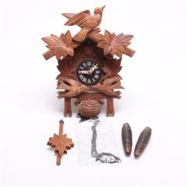 Brown Wood and Resin Cuckoo Clock: A brown wood and resin cuckoo clock. The clock has a house design with carved wood leaf and bird attachments. Other features include a carved wood frame and black finished face with raised white Roman numerals and pierced hands, a wood leaf pendulum and carved pine cone weights.
