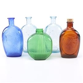Embossed Glass Bottles: An assortment of five embossed glass bottles. This assortment includes two light blue bottles, a blue bottle, a “Log Cabin” brown syrup bottle, and a green bottle. Some pieces are marked.