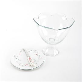 Glass Compote Bowl With Pfaltzgraff Plate and Knife: A glass compote bowl and a cheese plate and knife from Pfaltzgraff. This lot features a cheese plate with holly designs, and a cheese knife with textured holly and bell designs on the handle, and a serrated blade with a curved, double-pointed tip, marked “Stainless China”. Also included is a glass compote bowl with a floriform lip and subtle embossed designs.
