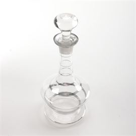 Czechoslovakian Glass Decanter: A glass decanter produced in Czechoslovakia. This decanter has a round stopper and a flared lip. It has a round neck which tapers outwards to join the body, and a round base. There are several silver tone bands running around the body, and the piece is marked to the underside “Czechoslovakia”.