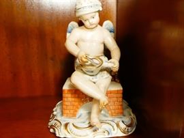 Meissen figurine representing a cherub sitting on a brick stove making porridge he wears a drape around his lower torso with a matching nightcap on his head late 19th century, hard-paste porcelain polychrome detail