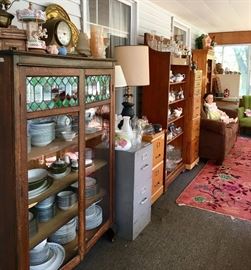 In the foreground is a beautiful, antique cabinet with intricate glass patterns in green and clear across the top shelf. Inside are sets of china dishes and on the top are a mantel clock, lamp shades, a lamp, etc.