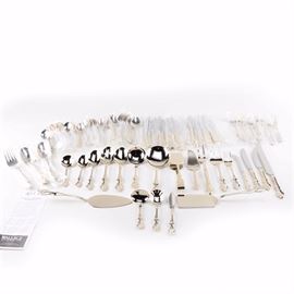 Wallace Silversmiths Stainless Steel Flatware Set: A set of stainless steel flatware by Wallace Silversmiths. This seventy-four piece collection includes twelve dinner forks, twelve dinner knives, twelve steak knives, twelve teaspoons, twelve round soup spoons, three tablespoons, three pieced tablespoons, three serving forks, two casserole spoons, two pie servers, a spatula, a meat carving set, a pair of tongs, a ladle, a sugar spoon, a master butter, and a cheese pick. These pieces features a scrolled foliate pattern to the handles and each is marked “RSVP TG Indonesia”.