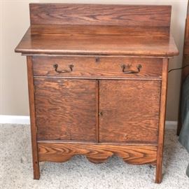 Antique Oak Washstand: An antique oak washstand. This piece is constructed from oak, and it features a rectangular top with backstop over one hand dovetailed drawer with metal drop handles. Below are double cabinet doors and a serpentine apron. This washstand rests on square-cut feet.