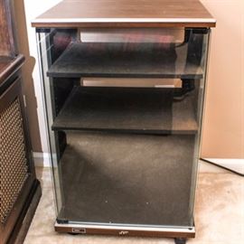 Vintage JVC Stereo Cabinet: A vintage JVC stereo cabinet. This piece is constructed from engineered wood with walnut and steel tone accents. It features a clear glass door, through which two shelves can be viewed, and it rolls on plastic casters.