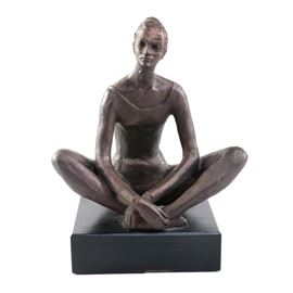 Nora Carol Resin Dancer Sculpture: A Resin sculpture of a dancer. This figural sculpture depicts a female dancer reflecting while she sits, stretching. This piece is signed “Nora Carol” to the underside of the dancer’s leg. The sculpture is presented atop a black, square base.