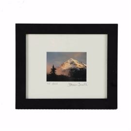 Steve Terrill Color Photograph "Mt. Hood": A color photograph on paper titled Mt. Hood by Steve Terrill. This piece captures the summit of Mt. Hood in Oregon at sunrise, with dazzling light hitting the snow cap, and trees in silhouette in the foreground. The work is signed and titled in pencil to the lower edge. This piece is presented under a white window mat in a black tone wood frame with hanging wire affixed to the verso.