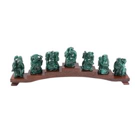Japanese Carved Malachite Seven Lucky Gods Netsuke Set: A set of seven Japanese carved malachite netsuke depicting the Seven Lucky Gods (七 福神 shichifukujin) with a wooden display stand. The figurines depict the Seven Lucky Gods including Ebisu, Daikokuten, Bishamonten, Benzaiten, Fukurokuji, Jurōjin, Hotei, and Kichijōten. Each has a double hole to thread a cord but are otherwise unmarked. They are presented with a stepped and footed wooden display stand.