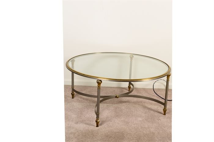 Glass Top Coffee Table: A glass top coffee table. This table features a circular top with brass tone trim and silver tone edges. It rises on four columnar legs with brass tone caps tops and tapered feet. A silver tone curved x-stretcher connects them.