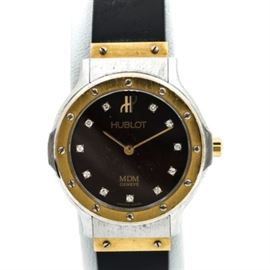 Hublot MDM Geneve Diamond Wristwatch: A Hublot MDM Geneve diamond wristwatch. This watch features a black rubber band leading to a silver and gold tone circular case housing a black dial with diamond hour indicators, slender gold tone hands, and imprinted with “Hublot MDM Geneve Swiss Made.”