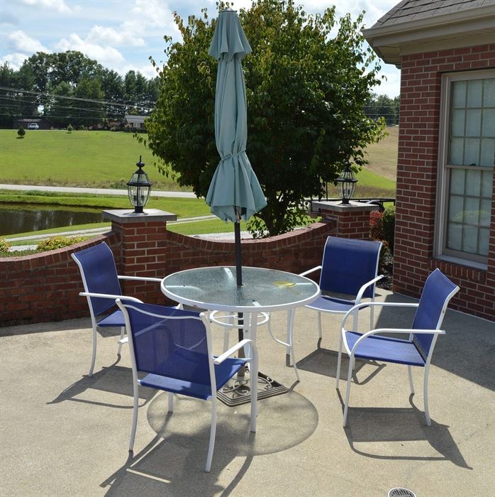 Patio Table with Umbrella and Four Chairs: A patio table with umbrella and four chairs. This includes one metal-framed table with a circular, hammered glass top; it rests on four splayed legs and its frame is painted white. The included umbrella has a light green shade and black pole. Each armchair features a white metal frame with a blue fabric seat and back.