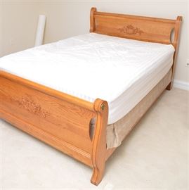 Victorian Style Oak Queen Size Sleigh Bed with Mattress: A Victorian style oak queen size sleigh bed with mattress. This bed frame features a headboard and footboard with scrolling crest rail and applied floral motif to the solid panel. The scrolling posts feature brass tone hardware to the top of the post edges. The boards are joined by side rails. There are no visible maker’s marks. Please note, the photographed bedding and mattress are not included.