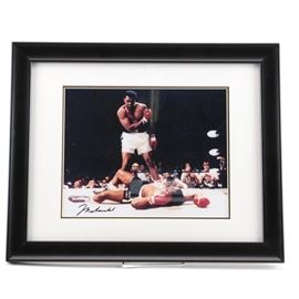 Autographed Muhammad Ali Photograph: An autographed Muhammad Ali Photograph depicting the iconic fight between Muhammad Ali vs. Sonny Liston in 1965. The photograph is autographed by Muhammad Ali in black ink to the bottom left. The images is set in a white mat and is presented behind glass in a black frame.