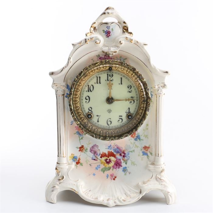 Victorian Style Royal Bonn Porcelain Case Mantel Clock: A Victorian Style Royal Bonn porcelain case mantel clock. This clock features a white porcelain case with with hand painted pansies, and floral motifs with gilt accents with ornate embellishments. The ivory clock face features black Arabic numbers, brass toned hands, and ornate brass bezel that opens on a hinge. The verso is open, and offers a set of three keys.