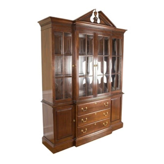 Ethan Allen Chippendale Style Breakfront China Cabinet: An Ethan Allen Chippendale style breakfront china cabinet. This cabinet features: a broken pediment with an urn style finial, wide molding with dentil detail, two doors on the upper section of the breakfront and single doors to either side all with two-over-three paned glass, three stacked drawers on the lower section of the breakfront with single paneled cabinet doors to either side, and a wide molding with a graduated edge. The sides are solid panels on both the upper and lower sections. Brass tone hardware on the drawers and doors accents the dark tone wood. Companions 17BAL110-133;17BAL110-135.