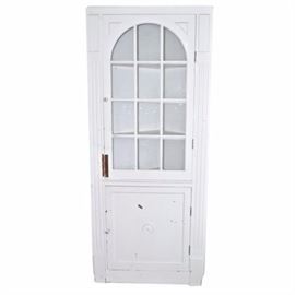 Shabby Chic Style Vintage Corner Cabinet: A Shabby Chic style vintage corner cabinet. The cabinet features reeded side columns and a top glass panel door with a rounded arch. It opens to reveal three storage shelves inside. The lower portion of the cabinet includes a wood door that opens to reveal one storage shelf. Each door includes white porcelain knobs with a painted floral motif. Please note, this item is located on a second floor.