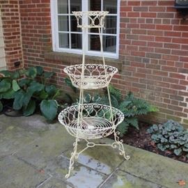 Metal Tiered Stand: A metal tiered stand. The stand has a white finish and features a three tiered design with three scroll work metal baskets in descending sizes displayed on a dowel frame with a three pole and ring stretcher with decorative scrolled legs and feet.