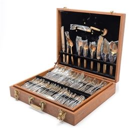 Bronze Tone Flatware Service for Twelve: A bronze tone flatware service for twelve featuring stylized anthemion motifs to the handle with beaded accents. The one hundred and forty-three piece service includes twelve each of dinner knives, serrated tip knives, butter knives, dinner forks, seafood forks, salad forks, appetizer forks, teaspoons, soup spoons and demitasse spoons. There are eleven long handle iced tea spoons and a matching twelve piece accessory grouping. The flatware is marked “Thailand SN” to handle verso and comes housed in a wooden flatware storage case with divided interior.