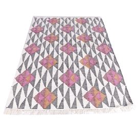 Handwoven Indian Wool and Cotton Area Rug: A handwoven Indian area rug. This wool and cotton rug features a geometric pattern of diamonds and triangles in deep charcoal, ivory, pink, orange, and crimson. It has overcast selvedges, finishing at each end with ivory cotton warp fringe. The rug is labeled on the reverse.