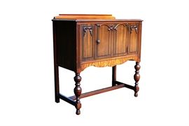 Art Nouveau Small Sideboard Cabinet: A small mahogany sideboard cabinet. It has a beveled back guard on the top, a single cabinet to the front, and is raised on vase and ring turned front legs joined to the block back legs by an H-stretcher. The front has four incised panels, with applied scrolled and arched leaf accents along the top. The center two panels form the solid door, which has a rosette knob and opens to storage spanning the entirety of the cabinet, with cord hole to one corner; the outer two are accented with an incised center arch. Down the front stiles are fluted accents, and the scalloped apron has a curly veneer.