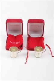 Reed & Barton Sterling Silver Annual Baby Cup Christmas Ornaments: A pair of sterling silver annual Christmas ornaments by Reed & Barton. Each cup features a personal engraving to the front reading, “Baby’s First Christmas 2008” and a scrolled foliate repoussé shield to the back. There is a sterling loop to each handle with a red ribbon attached. The underside of each is marked “Reed & Barton Sterling”. Each ornament comes in a red Reed & Barton snap jewelry boxes with red pouches. Total approximate weight: 1.03 ozt.