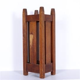 Antique Oak Mission Style Umbrella Rack by Stickley Bros., Circa Early 1900s: An antique Stickley Brothers umbrella stand from the Quaint Furniture line, circa early 1900s. This dark stained oak Mission style piece has a square base raised on post legs at each corner, cut to pyramidal points at the top. It has a panel border around the top and bottom, with the legs stretching above and below it, set with a solid panel in the center of each side, leaving open vertical slats. It has a metal drip tray in the bottom. On the base is a metal tag with “Quaint Furniture/ Stickley Bros./ Grand Rapids, Mich” to the right of the early logo.
