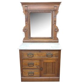 Eastlake Style Marble Top Washstand with Mirror: An Eastlake style marble top washstand with mirror. This piece is walnut with a swivel mirror and a white marble top. The mirror is within a frame with crown molding to the top, carved detail to the sides and incised lines and pressed rosettes. The washstand has a rectangular white marble top with gray veining. Below is a full-width drawer above two half-width drawers and a recessed panel cabinet door. The drawers have pin and crescent joinery, and ornate brass bail handles with floral wreath accented backplates. The chest stands on a plinth base.