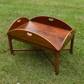 Mahogany Butler Tray Coffee Table by Henredon: A mahogany coffee table by Henredon. This butler’s tray style table has a rectangular center with bookmatched flame veneer, with a semicircular leaf to each side. The leaves flip up to resemble a butler’s tray, with a cutout handle in the center of each arch, or down to make the table’s top oval. It is raised on block legs connected with a cross stretcher. To the bottom, it is branded with the Heritage Henredon logo, retains a Mahogany Association tag, and is stamped “CI-II 7-53.”