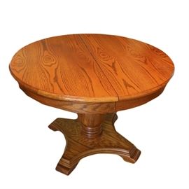 Oak Pedestal Dining Table: An oak pedestal dining table. The circular top splits in the center for an extension leaf. It has a central turned pedestal on square base with concave curved, beveled sides.