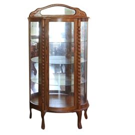 Vintage Oak Bow Front Curio Cabinet: A vintage oak bow front curio cabinet. This demilune top oak cabinet with serpentine back splat features a mirrored panel and applied scroll detail. The cabinet features a central full length, clear glass panel door opening to three glass shelves, a mirrored back, and recessed lighting. The cabinet door is flanked by turned stiles and glass side panels. The cabinet rises on short cabriole legs and terminates on claw feet. There are no visible maker’s marks. For a matching piece, see item 17DAL172-044.