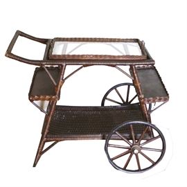 Vintage Wicker Bar Cart: A vintage wicker bar cart. This vintage bar cart features a wooden wicker wrapped frame, large handle, rectangular glass removable tray top framed wicker dual handled frame, side shelves and a bottom shelf. Cart has two large front spoke style wheels and straight legs to the back.