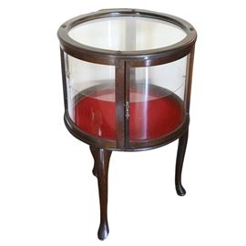 Queen Anne Style Curio Drum Table: A Queen Anne style curio table. It has a dark stained wooden frame on three cabriole legs with pad feet. The top and sides are glass paned, and a single side door opens to a glass shelf and a red felt lined base.