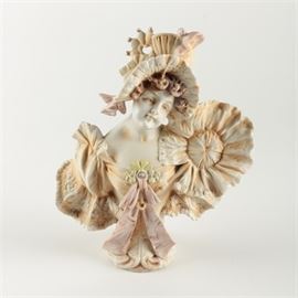 Porcelain Bust of a Woman: A porcelain bust of a woman. The woman wears a large, decorated hat with ribbons and flowers matching her floral and ruffled dress with ribbon accents. She has curled red hair and minimal coloring throughout. The bust is marked “JPK 1690” to the underside.
