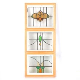 Arts and Crafts Style Stained Glass Windows in Wooden Frame: A set of three stacked stained glass windows in a wood frame. The Arts and Crafts style windows each feature multiple panes of glass with stained glass designs to the center of each window. The pieces feature highlight colors of amber, burgundy and lime. Each of the original frames was painted in an ivory or cream tone, and the windows are stacked together in a natural finish wooden frame.