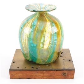 Blown Glass Vase on Wooden Stand: A blown glass vase on a wooden stand. The vase has a flared lip with a short neck and a wide body. The vase has a swirled green and yellow design and is presented on a wooden stand.