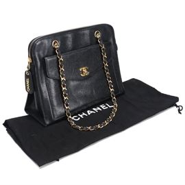 Chanel Front Pocket Tote: A Chanel tote in black caviar leather. This classic bag has a large flap pocket on the front secured with a gold tone logo turn lock. Two leather woven chain straps are present to the top. A main zipper closure with a statement Chanel pull opens to reveal a black textile lined interior with a single zippered sidewall pocket and a leather brand emblem. Accompanying the purse is a black Chanel dust cover.