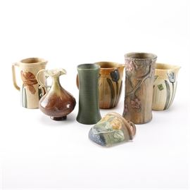 Art Pottery Including Roseville Sunflower and Grueby Faience: A selection of Art Pottery including Weller and Grueby. Included is a simple Grueby Faience green matte vase, showing the maker’s throwing rings to the exterior and having a pale green, slightly crazed liner glaze. It is stamped to the underside with the circular “Grueby Faience Co. / Boston U.S.A.” mark to the underside, and a production number or date. This piece includes minor damage-chipping-to the bottom rim. Also included is a Weller vase in the Woodcraf pattern, circa late 1910s to mid 1920s, having a striated soft amber brown matte glaze with a pattern of branches and pink blossoms to the exterior and a pale honey amber liner glaze. Marked to the underside with he impressed “Weller” name in serif font. Included is a Roseville ewer in the Rozane pattern, having an R.P. Co impressed mark with a pattern number, in a ruffled rim form with decorative handle. Included is a Roseville Sunflower wall pocket, having a sunflowe